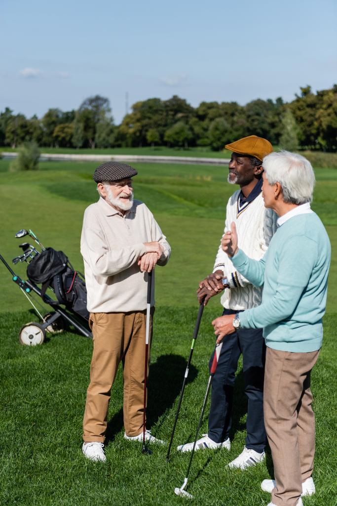 senior multiethnic friends smiling and holding golf clubs on green lawn - Фото, изображение