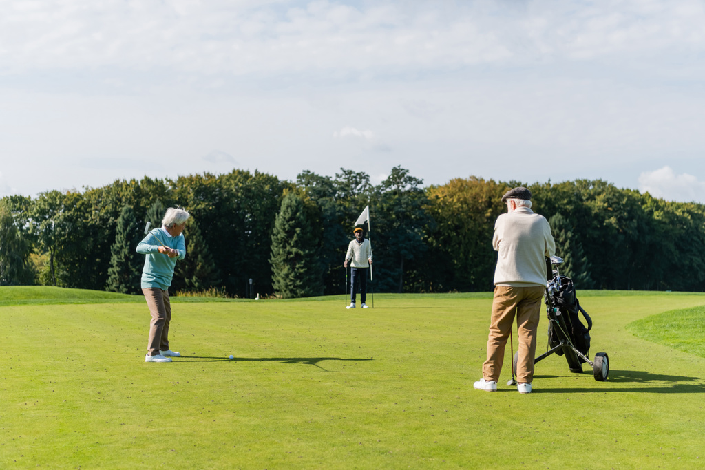 старший азіатський чоловік грає в гольф біля міжрасових друзів з прапором палицею і кошиком для гольфу
 - Фото, зображення