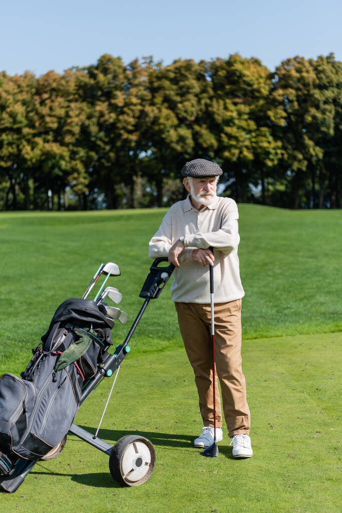 bebaarde senior man met platte cap staande in de buurt van golfkar  - Foto, afbeelding