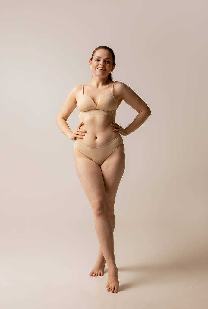 Full-length portret van jonge mooie vrouw poseren in beige ondergoed geïsoleerd over studio achtergrond - Foto, afbeelding