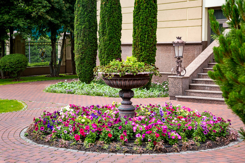Stenen bloem bed fontein in de achtertuin met bloeiende petunia 's met een bakstenen tegel bestrating in de voorkant van een granieten gebouw en een ijzeren lantaarn straatverlichting, niemand. - Foto, afbeelding