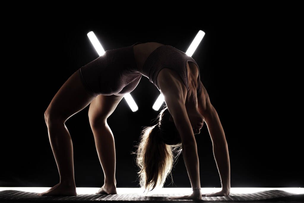 Fit Frau praktiziert Yoga-Posen. Silhouette Mädchen bei Übungen im Studio vor schwarzem Hintergrund mit v-förmigem weißen LED-Röhrenlicht. Kein Konzept zum inneren Ausgleich. - Foto, Bild