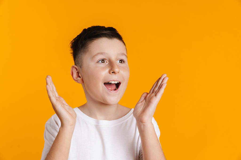 szczęśliwy uśmiechnięty podekscytowany nastolatek chłopiec w t-shirt stojąc na żółtym tle trzymając ramiona na twarzy - Zdjęcie, obraz
