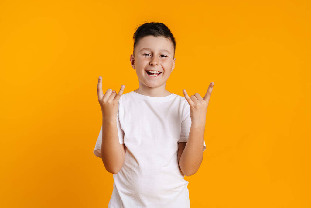 Brunette garçon blanc en t-shirt souriant et faisant geste de corne isolé sur fond jaune - Photo, image