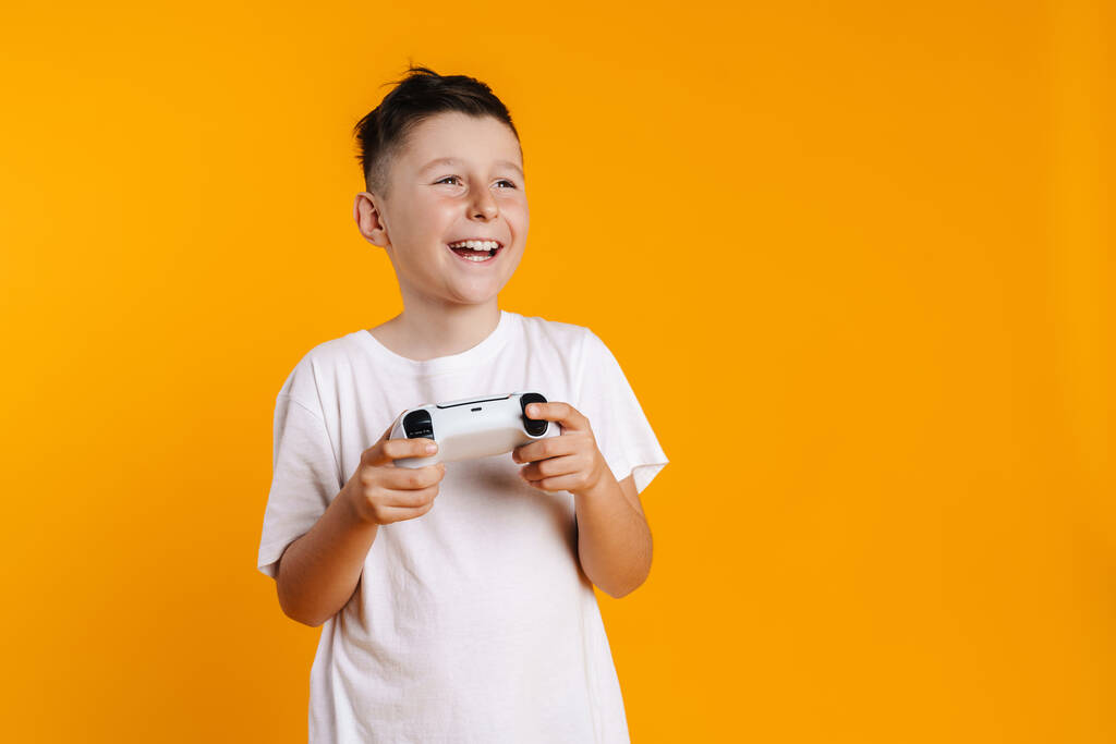 黄色の背景に隔離されたジョイスティックでオンラインゲームをプレイ中に笑顔ブルネットの白い少年 - 写真・画像