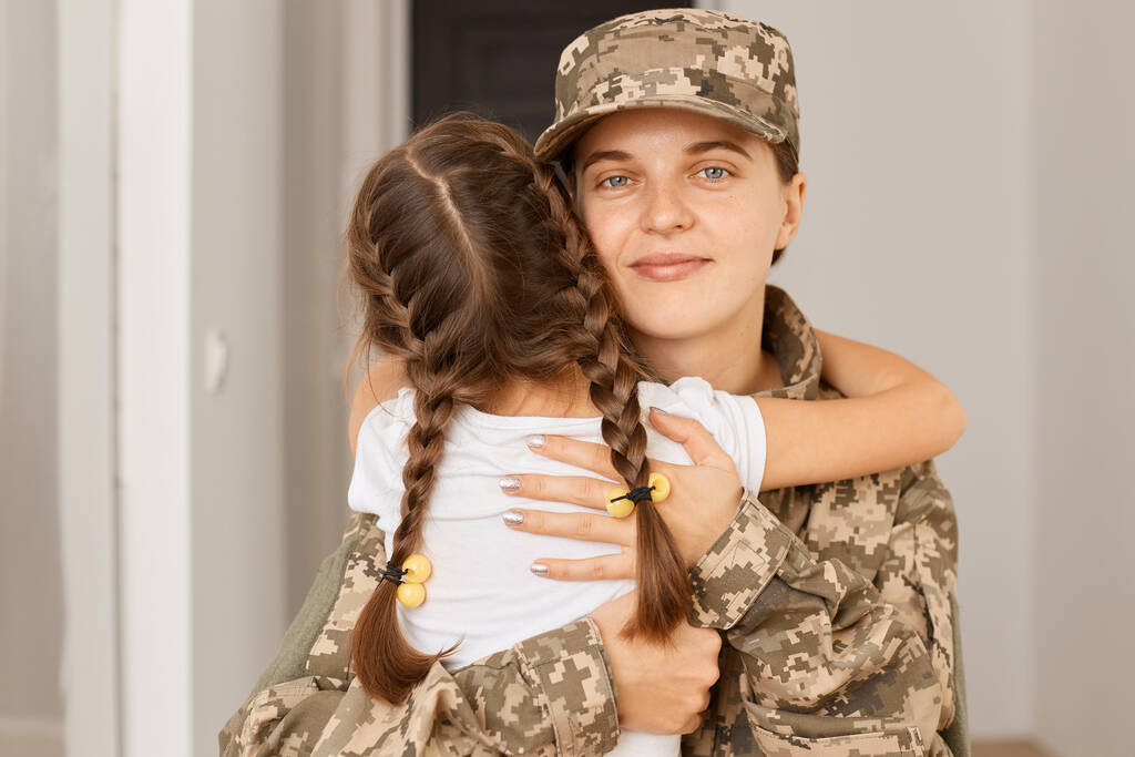 Portret uśmiechniętej, zachwycającej kobiety w mundurze kamuflażu i czapce pozującej z córką, przytulającej matkę, cieszącej się na jej widok po długiej separacji. - Zdjęcie, obraz