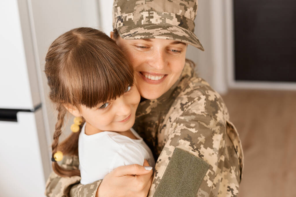 Portrait of smiling young adult soldier woman wearing camouflage uniform and cap posing with her daughter, hugging kid with charming smile, being happy to meet after being in army long time. - Photo, Image