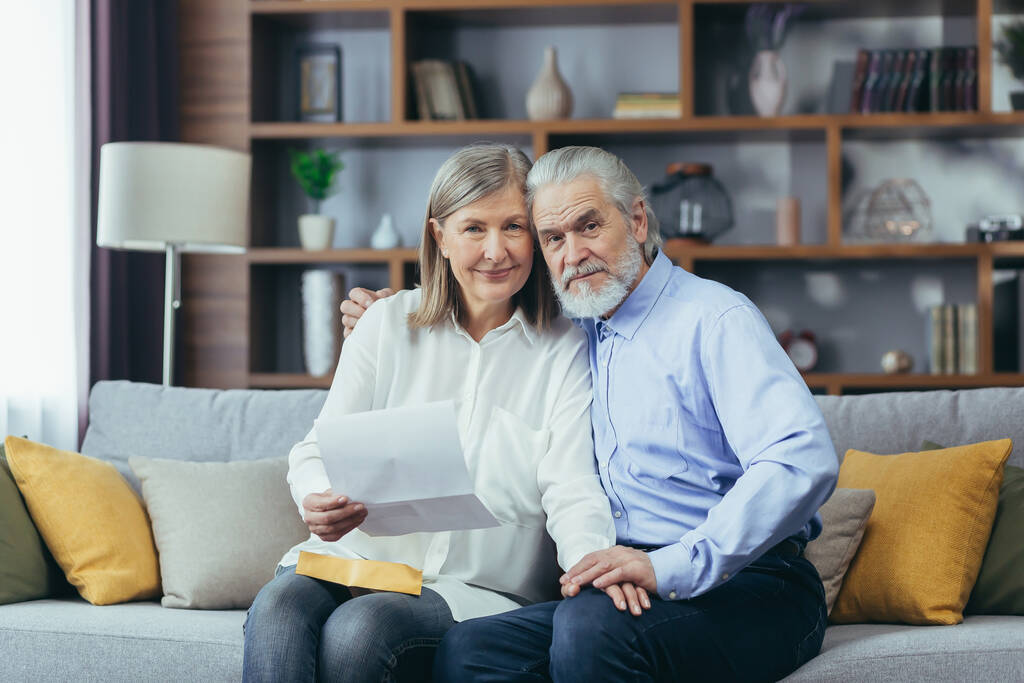 Senior couple man and woman together at home, received good news letter, happy and smiling sitting on sofa, looking at camera - Photo, Image