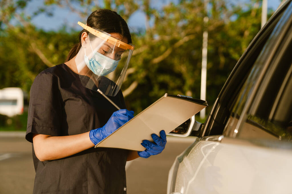 Trabajador de la salud negro en máscara facial y guantes anotando notas mientras está de pie en el coche al aire libre - Foto, Imagen