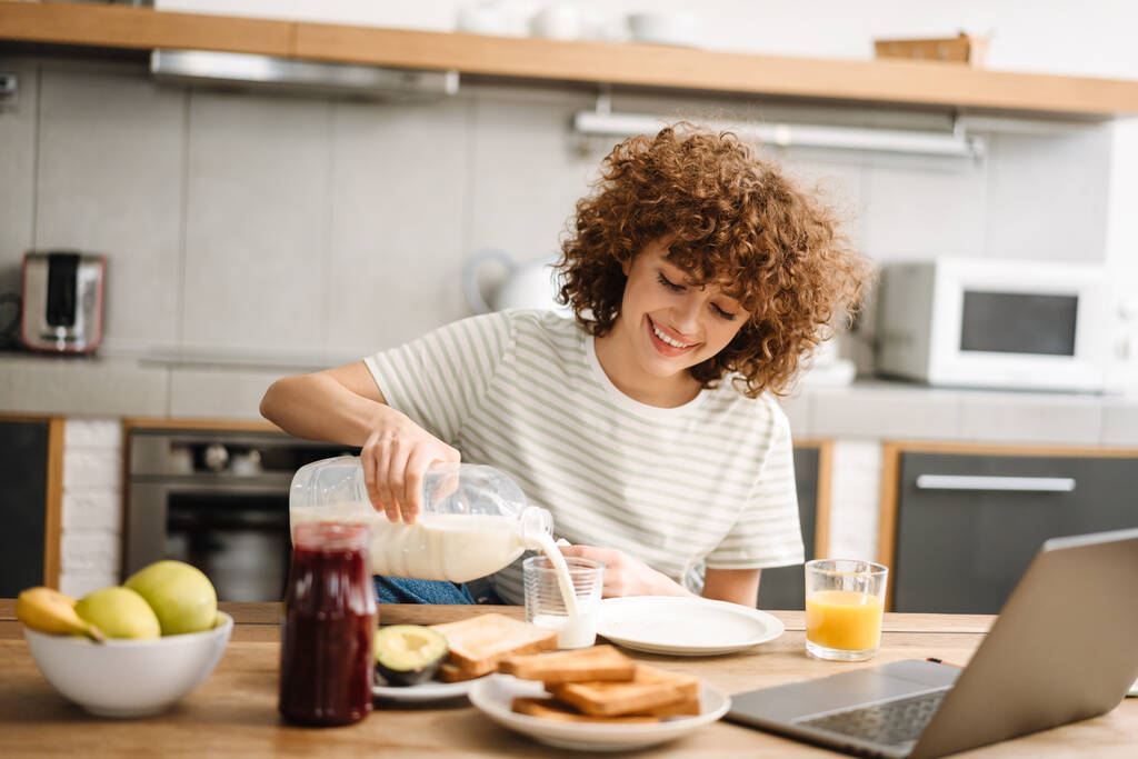 Улыбающаяся молодая женщина завтракает, сидя за столом на кухне - Фото, изображение