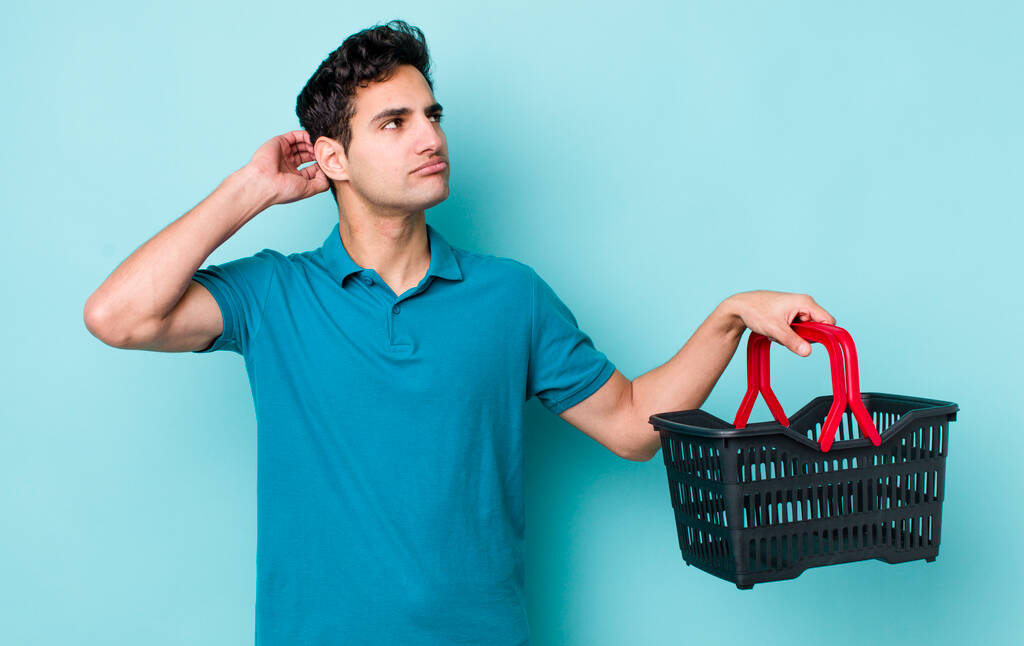 Homem hispânico bonito sentindo-se intrigado e confuso, coçando a cabeça. conceito de carrinho de compras - Foto, Imagem