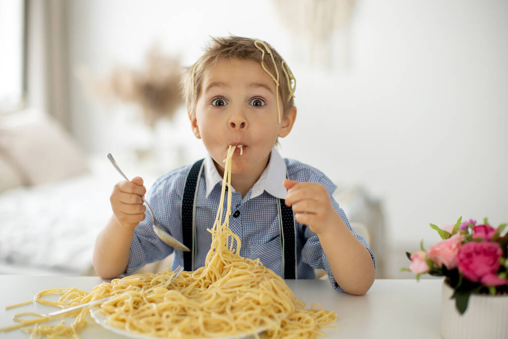 Χαριτωμένο παιδί προσχολικής ηλικίας, ξανθό αγόρι, τρώει μακαρόνια στο σπίτι, κάνει ένα χάος παντού, αστείες στιγμές - Φωτογραφία, εικόνα