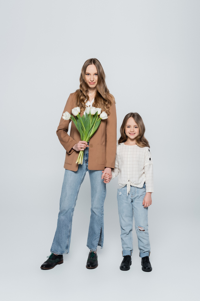 plná délka pohled stylové ženy s tulipány držící se za ruce s dcerou na šedém pozadí - Fotografie, Obrázek