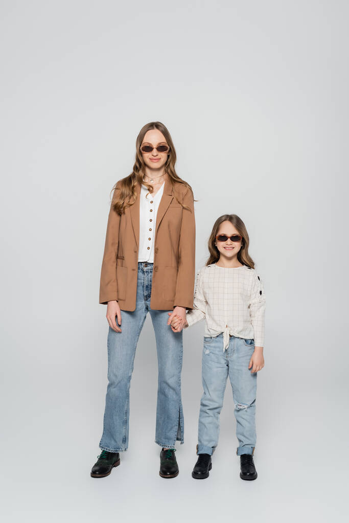 Вид в полный рост трезвенных матери и дочери в солнцезащитных очках, держащих руки на сером фоне - Фото, изображение