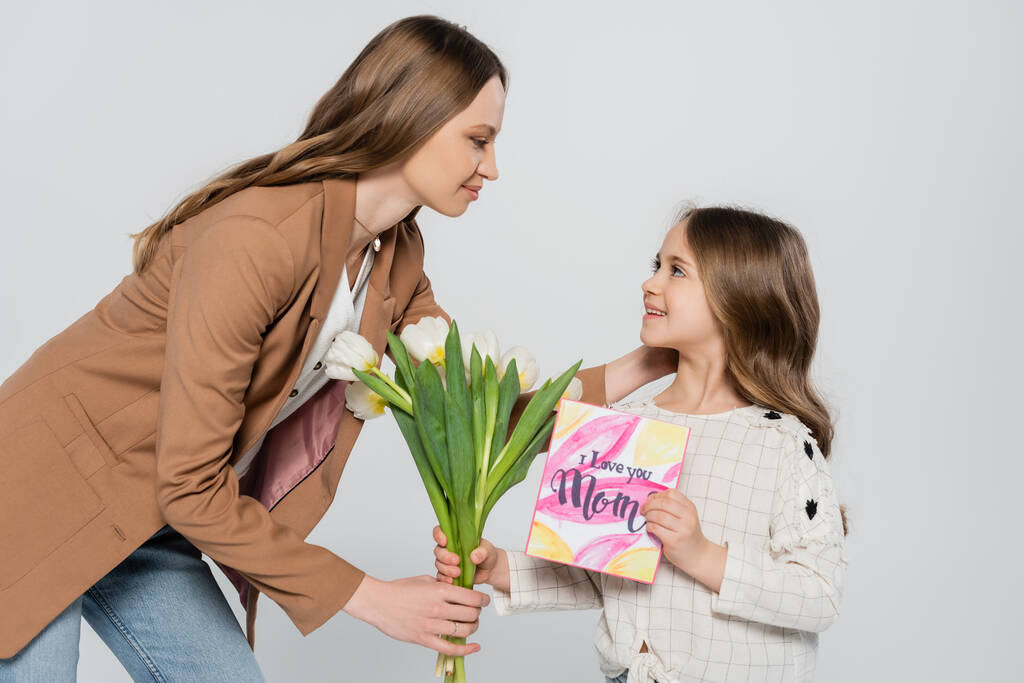 fröhliches Mädchen überreicht Tulpen und Grußkarte an Mutter isoliert auf grau - Foto, Bild