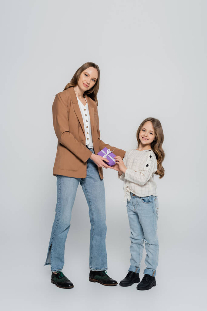 full length view of trendy mother and daughter smiling near gift box on grey background - Photo, Image