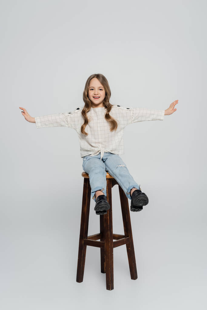 plná délka pohled na veselý dívka sedí na vysoké stoličce s natažené ruce na šedém pozadí - Fotografie, Obrázek
