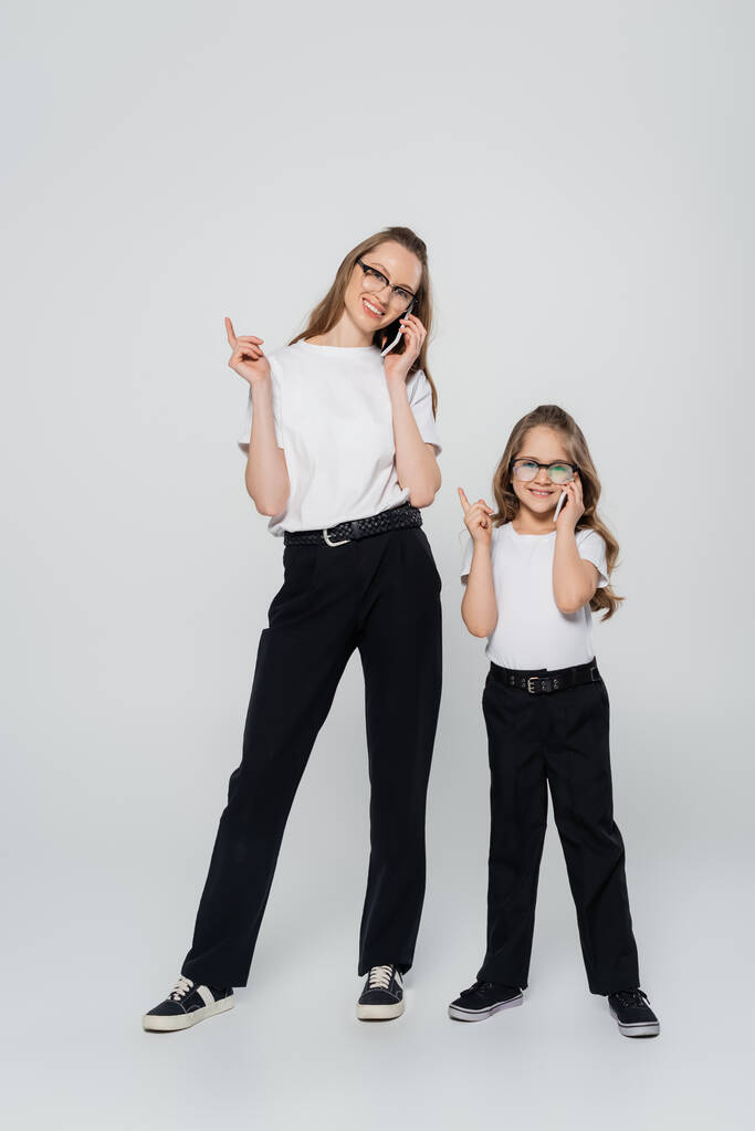 full length view of mother and daughter talking on cellphones and pointing with fingers on grey background - Photo, Image