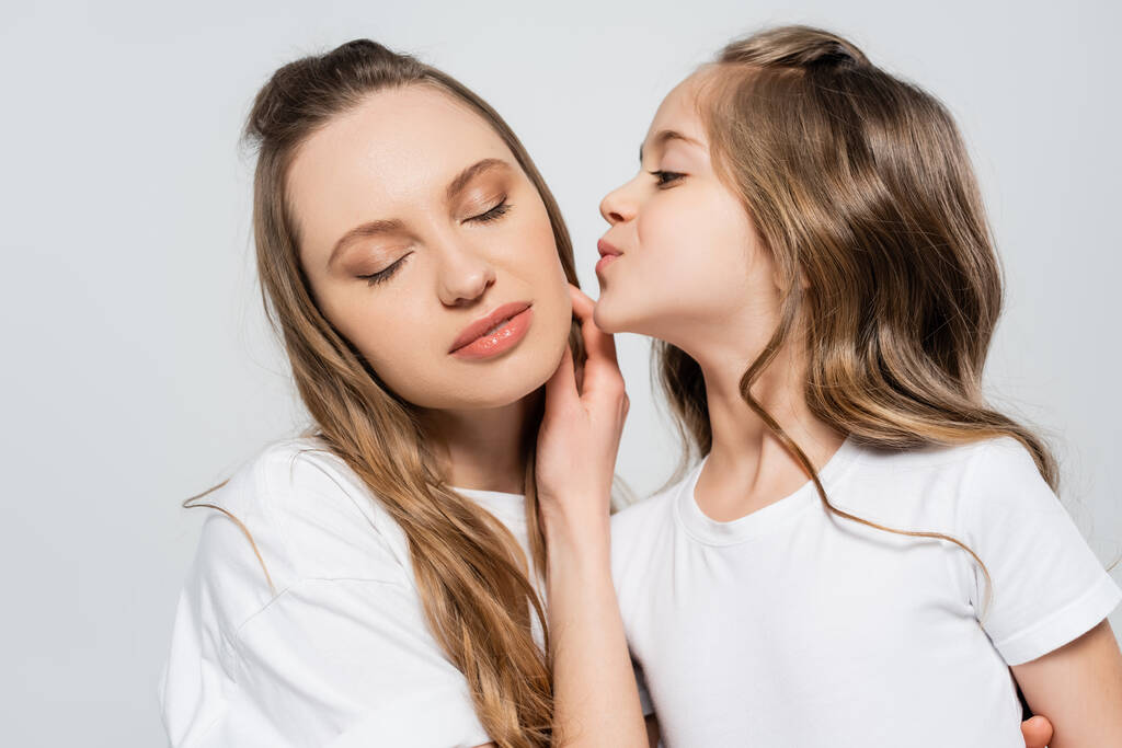 Frau mit geschlossenen Augen nahe Tochter küsst sie isoliert auf grau - Foto, Bild