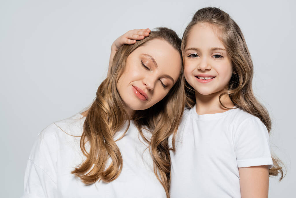 cheerful girl looking at camera near mother with closed eyes isolated on grey - Photo, Image