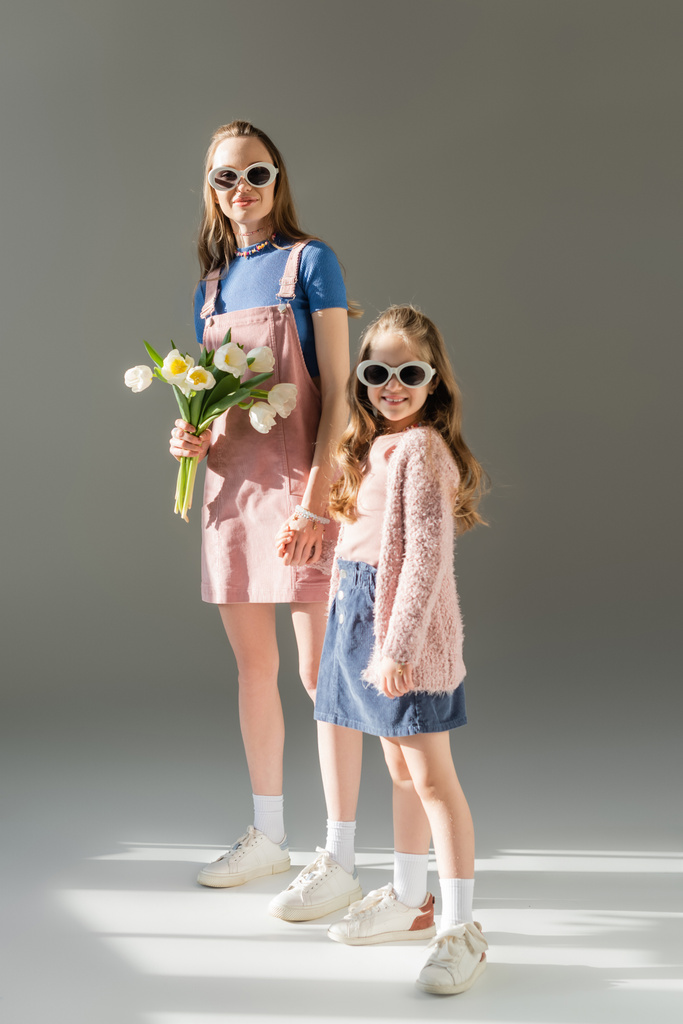 повна довжина щасливої матері з квітами і дочкою в сонцезахисних окулярах тримає руки на сірому
  - Фото, зображення