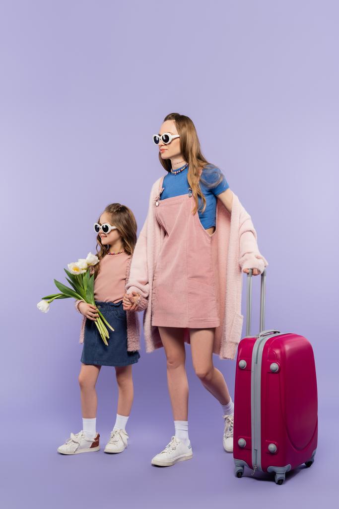 Mutter und Kind in Sonnenbrille halten sich an Händen und gehen mit Gepäck auf lila - Foto, Bild