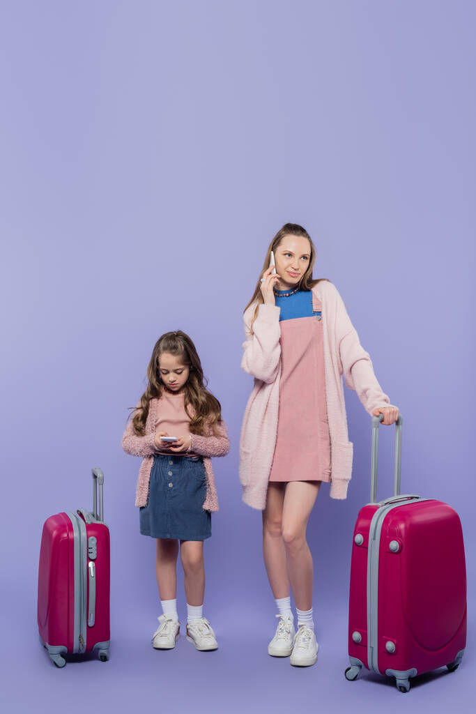 πλήρες μήκος της μητέρας και του παιδιού χρησιμοποιώντας smartphones κοντά στις αποσκευές σε μωβ - Φωτογραφία, εικόνα