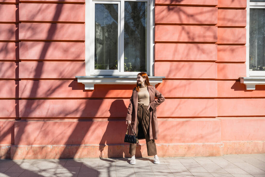 piena lunghezza della donna rossa allegra in piedi cappotto con borsa vicino edificio  - Foto, immagini