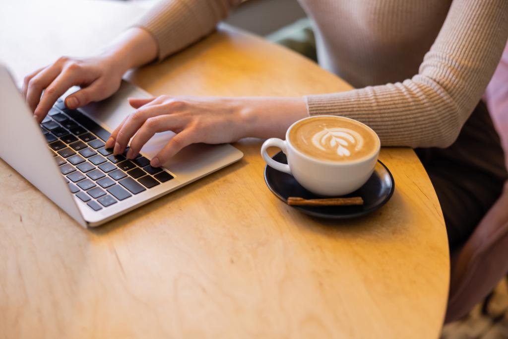 kivágott kilátás fiatal szabadúszó segítségével laptop közelében csésze cappuccino  - Fotó, kép