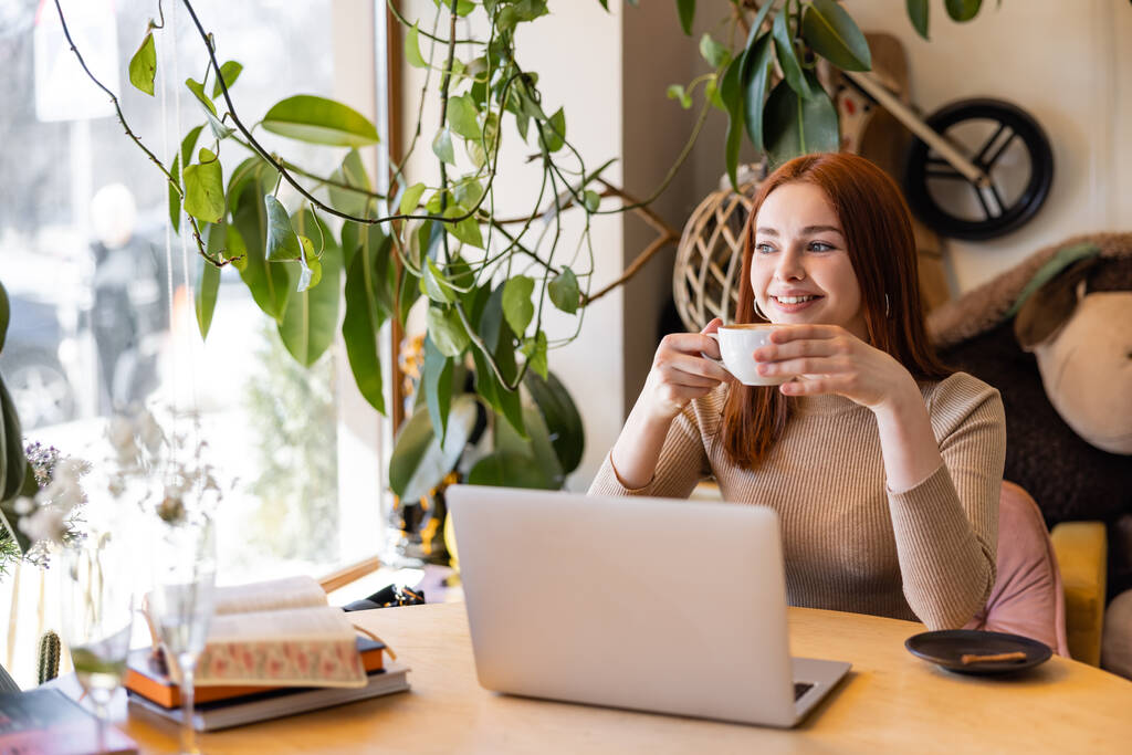 usmívající se žena s rudými vlasy drží šálek kávy v blízkosti notebooku v kavárně  - Fotografie, Obrázek