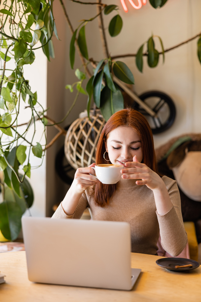 allegra giovane donna con i capelli rossi in possesso di tazza di caffè vicino al computer portatile in caffè  - Foto, immagini