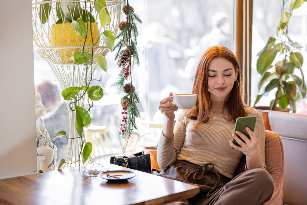 zadowolona młoda kobieta z rudymi włosami za pomocą smartfona i trzymając filiżankę kawy - Zdjęcie, obraz