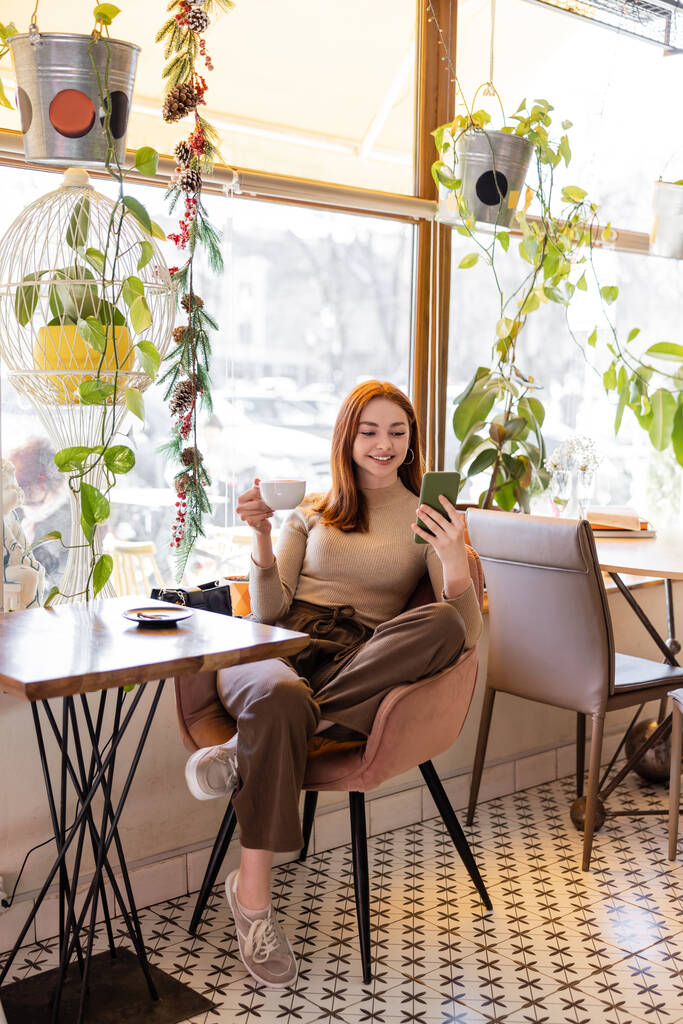 örömteli fiatal nő vörös haj segítségével okostelefon és gazdaság csésze kávé az asztalon  - Fotó, kép