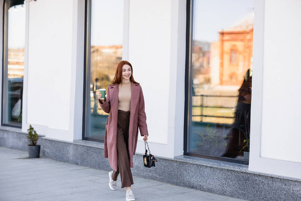 full length of pleased redhead woman with handbag holding paper cup and walking on urban street  - Photo, image