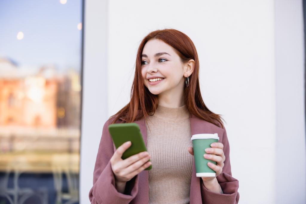 cheerful woman holding coffee to go and smartphone on street - Foto, imagen