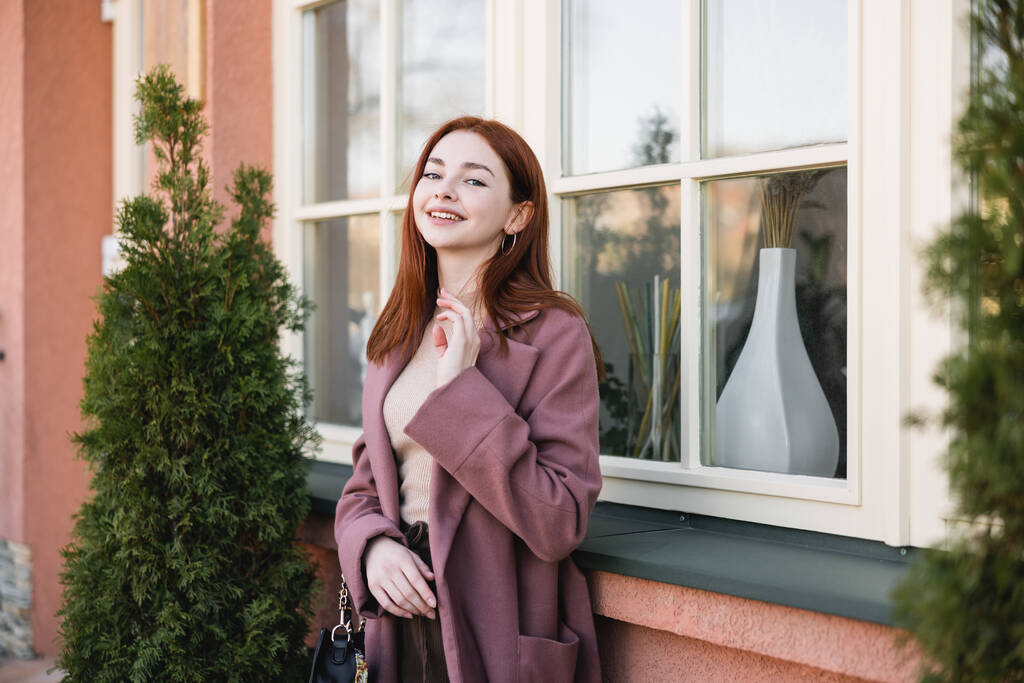 young cheerful woman in coat looking at camera near building  - Photo, Image