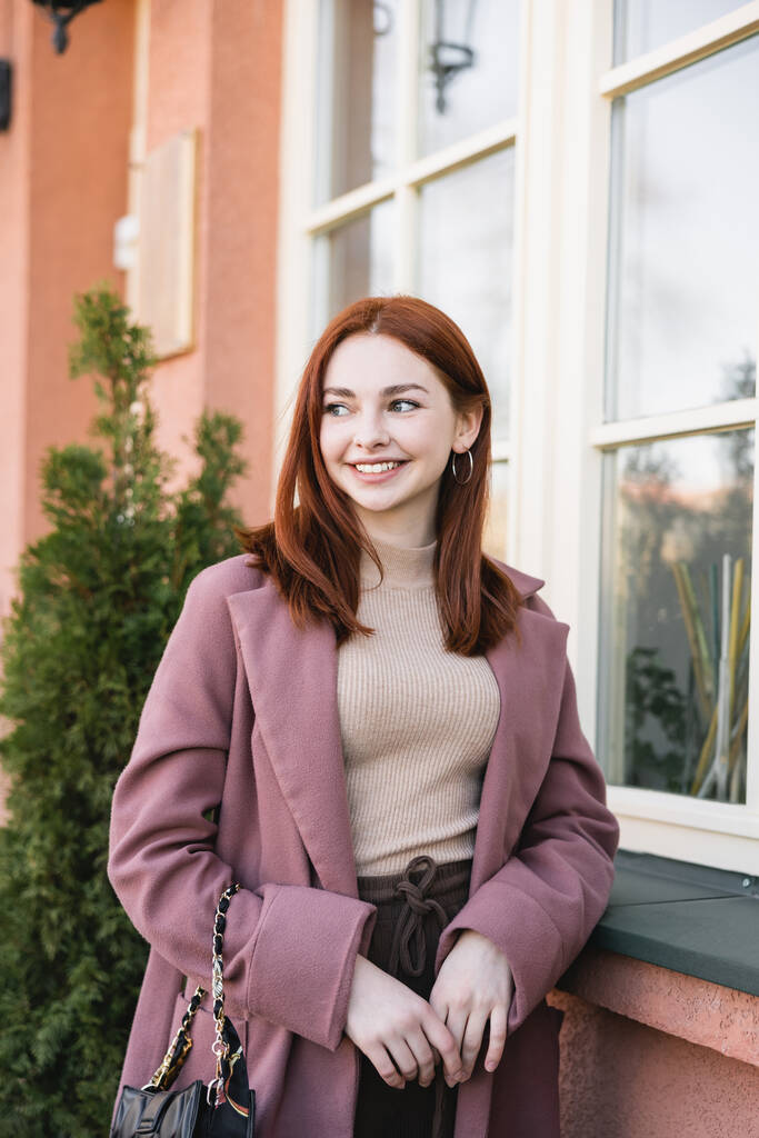 Nuori tyytyväinen nainen punaiset hiukset hymyilee lähellä rakennuksen ulkopuolella - Valokuva, kuva