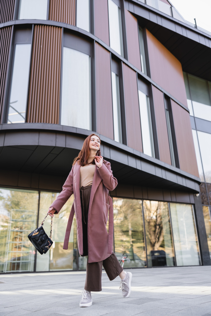 happy redhead woman in coat with handbag walking near modern building  - Foto, Imagen