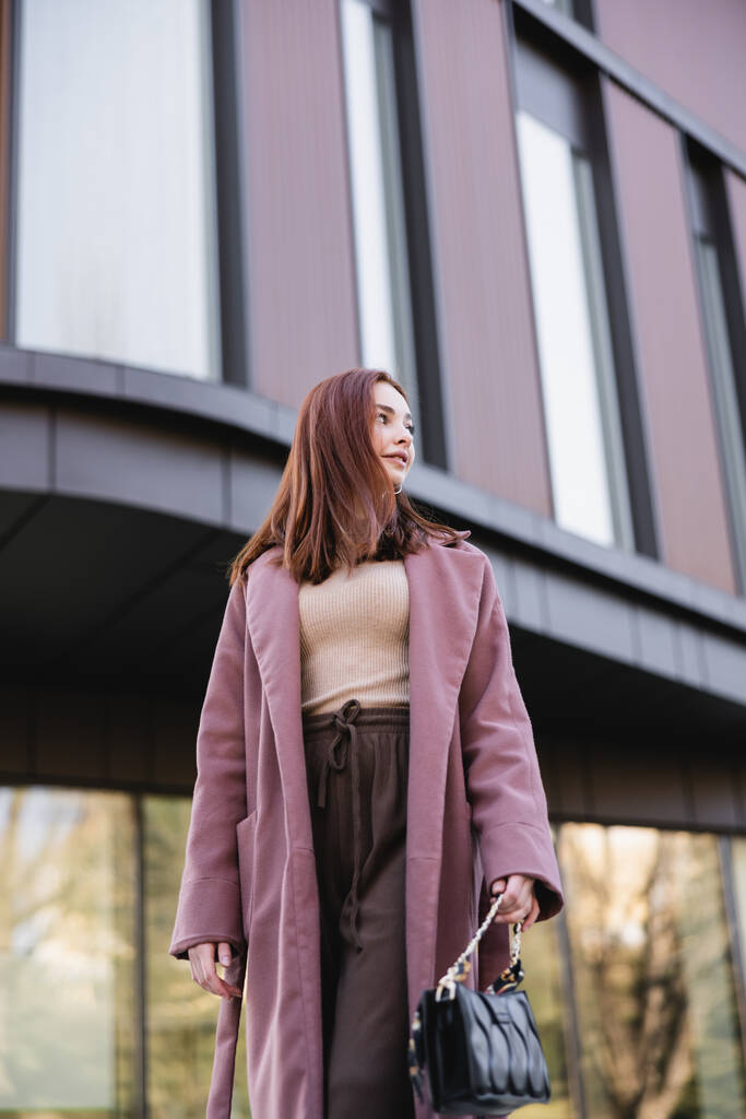 Modern binanın yanında duran el çantalı, paltolu genç kızıl saçlı kadının düşük açılı görüntüsü.  - Fotoğraf, Görsel