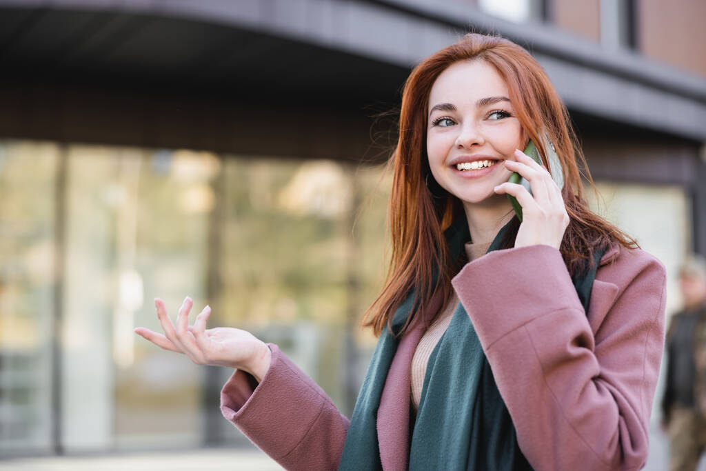 счастливая рыжая женщина в шарфе и пальто разговаривает на смартфоне снаружи  - Фото, изображение