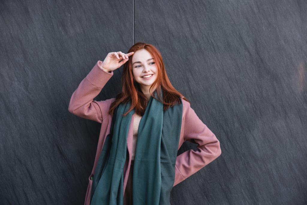 happy redhead woman in scarf and coat smiling near grey marble wall  - 写真・画像