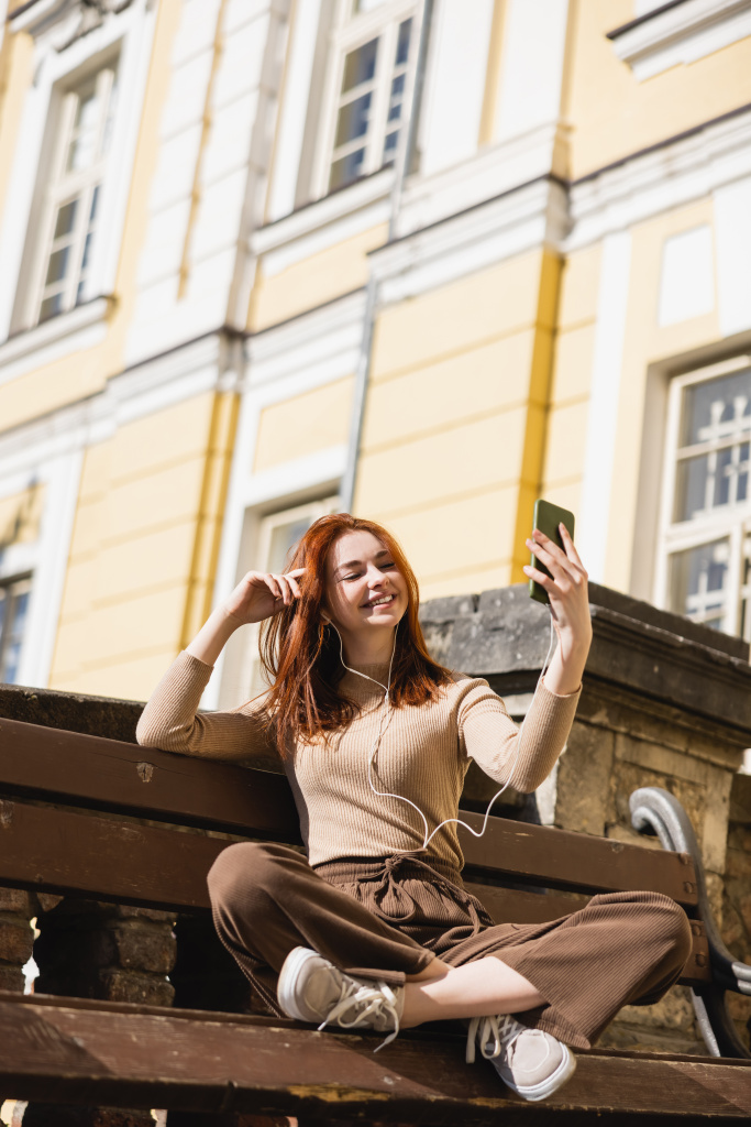 Frau hört Musik in kabelgebundenen Kopfhörern und macht Selfie im Sitzen auf Bank - Foto, Bild