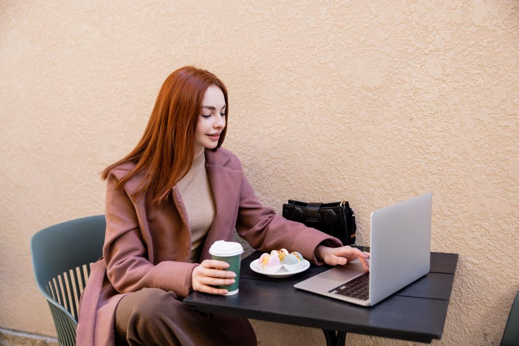 young redhead freelancer using laptop and holding coffee to go near plate with macarons  - 写真・画像