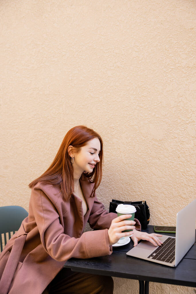 vidám nő kezében kávé menni, és használja laptop a kávézó teraszán  - Fotó, kép
