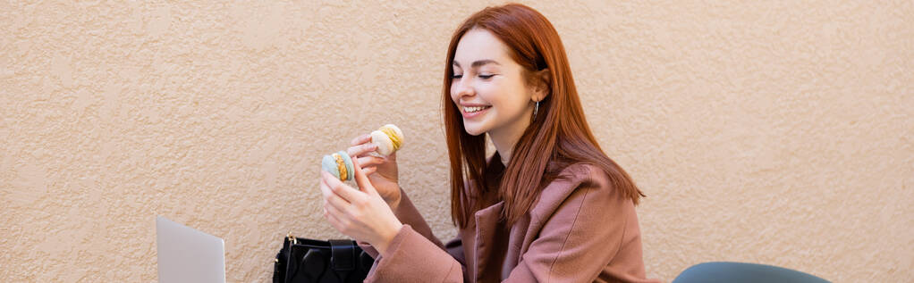 feliz joven con el pelo rojo sosteniendo macarrones dulces cerca de la computadora portátil en la terraza de la cafetería, pancarta - Foto, imagen