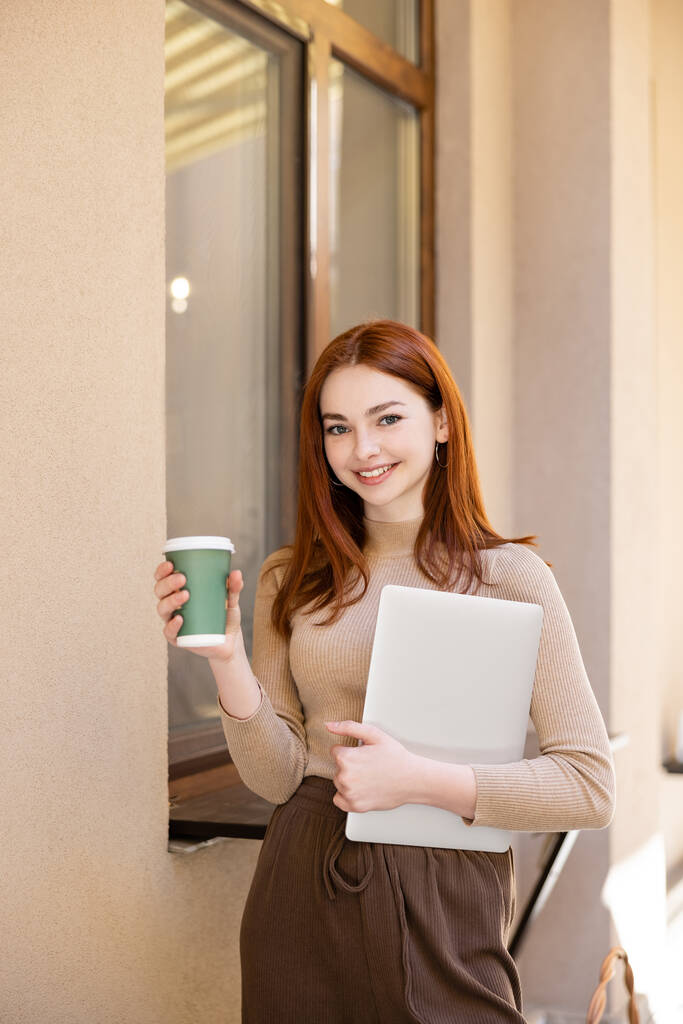 donna sorridente con i capelli rossi che tengono il computer portatile e tazza di carta vicino alla costruzione  - Foto, immagini