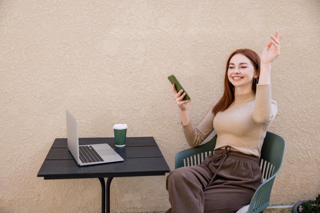 pelirroja freelancer en cuello alto sosteniendo smartphone mientras llama camarero en terraza de verano - Foto, imagen