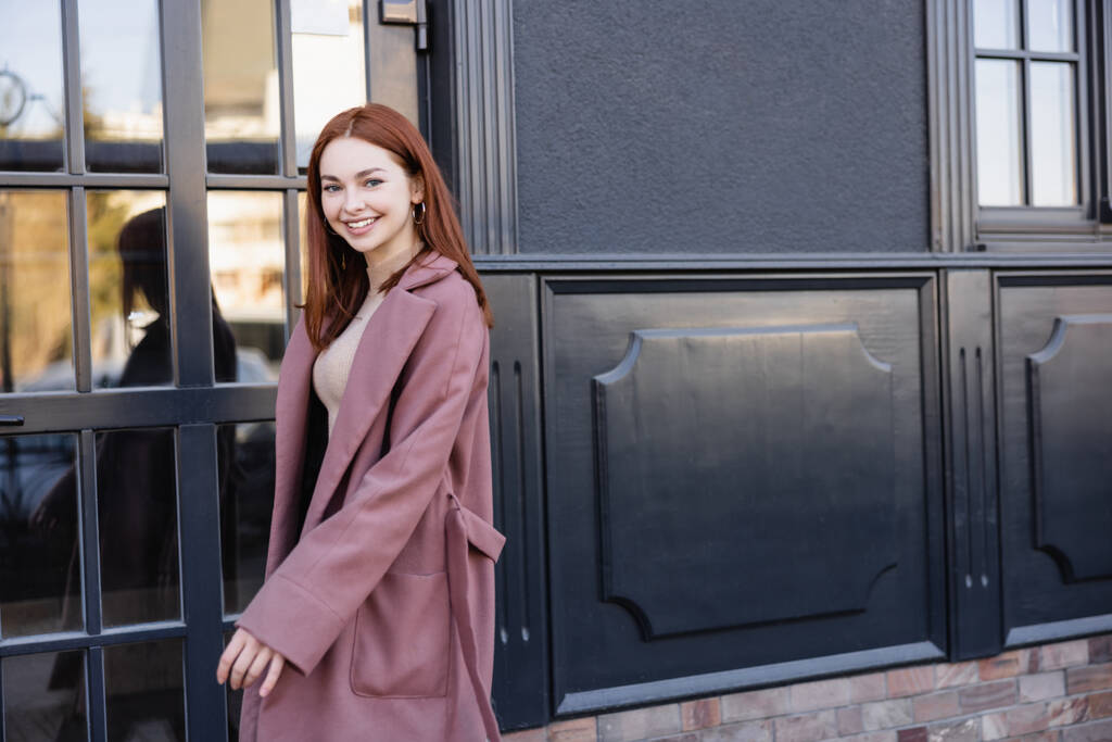 szczęśliwy ruda kobieta w stylowy płaszcz uśmiechnięty w pobliżu nowoczesny budynek  - Zdjęcie, obraz