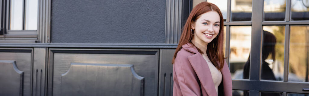 mulher ruiva feliz no casaco elegante sorrindo perto do edifício moderno, banner - Foto, Imagem