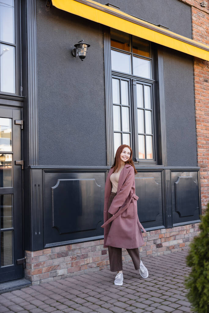 koko pituus onnellinen nainen tyylikäs takki kävely lähellä modernia rakennusta  - Valokuva, kuva